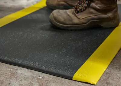 A close-up shot of work boots standing on top of a black safety mat with a bright yellow trim on either side of the mat. The mat is on a concrete floor.