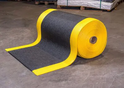 A role of black textured safety matting with yellow edges on a concrete floor. A broken pallet with plastic wrapped items is in the background.