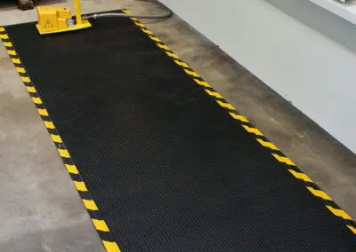 A black safety mat with a dotted surface and yellow and black hashing down one side in front of an industrial machine.