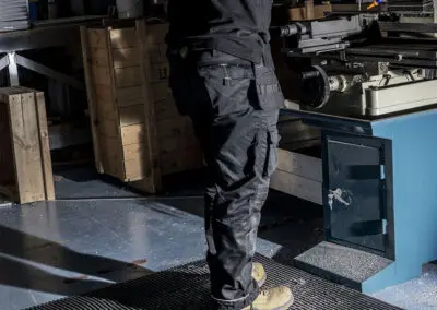 An engineer, dressed in all black clothing, using a drilling machine, while standing on a black textured mat.
