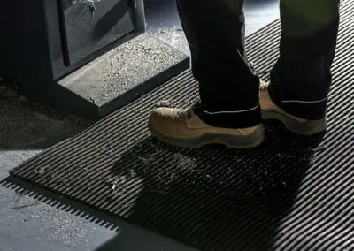 A close up of someone wearing a pair of brown working boots stood on a ribbed, black safety mat. There is metal shavings and swarf all over the mat and their boots.