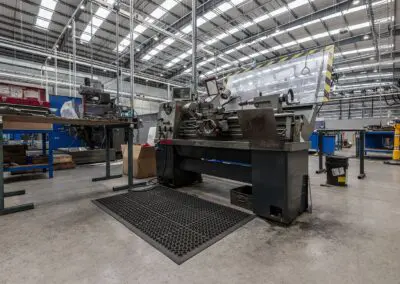 A safety mat with regular circular holes in it placed in front of a metalworking lathe in a workshop environment.